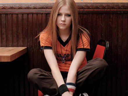 A high-definition desktop wallpaper featuring Avril Lavigne, seated with a serious expression, dressed in an orange music-themed shirt against a wooden background.