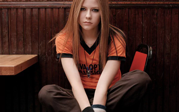 A high-definition desktop wallpaper featuring Avril Lavigne, seated with a serious expression, dressed in an orange music-themed shirt against a wooden background.