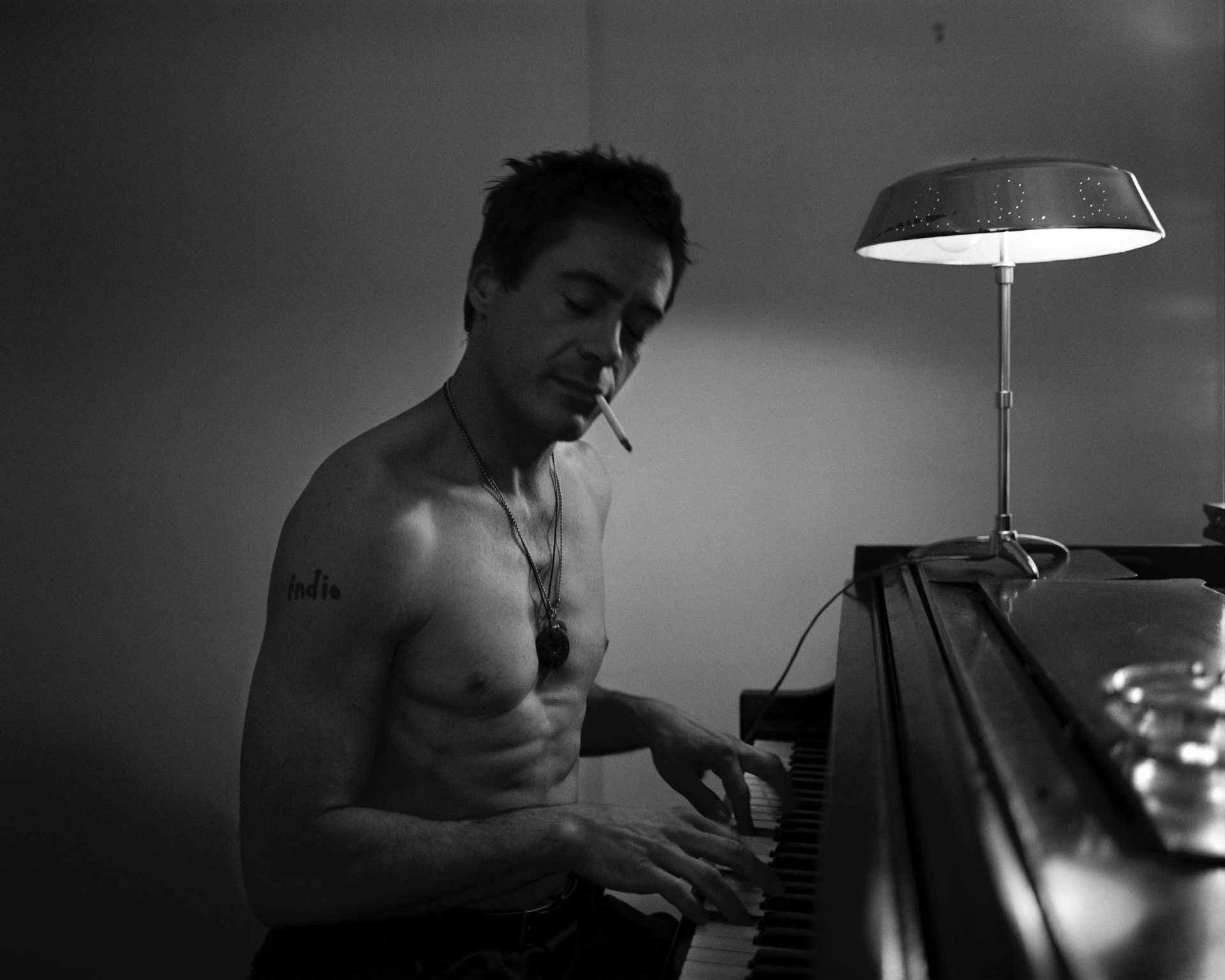 Black and white HD PC wallpaper of Robert Downey Jr. playing piano shirtless with a cigarette, illuminated by a desk lamp.