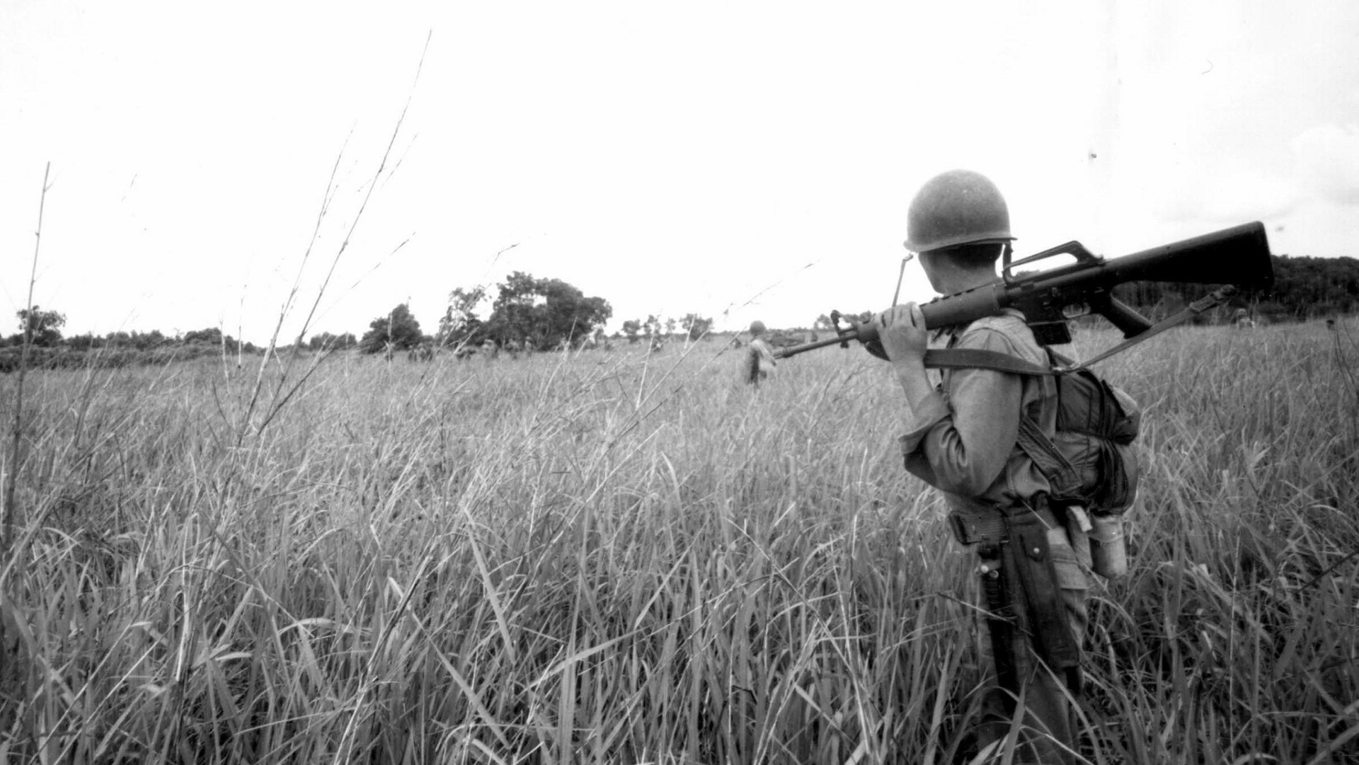 Black and white HD PC desktop wallpaper featuring a soldier in military gear carrying a weapon while walking through a tall grassy field.