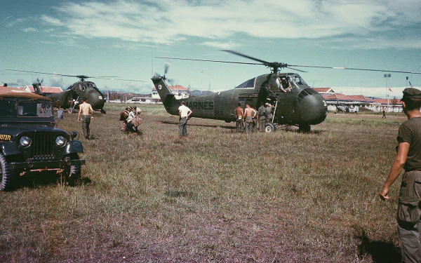 Military Sikorsky H-34 on a grassy airfield surrounded by crew, support vehicles and hangars — HD PC desktop wallpaper/background.