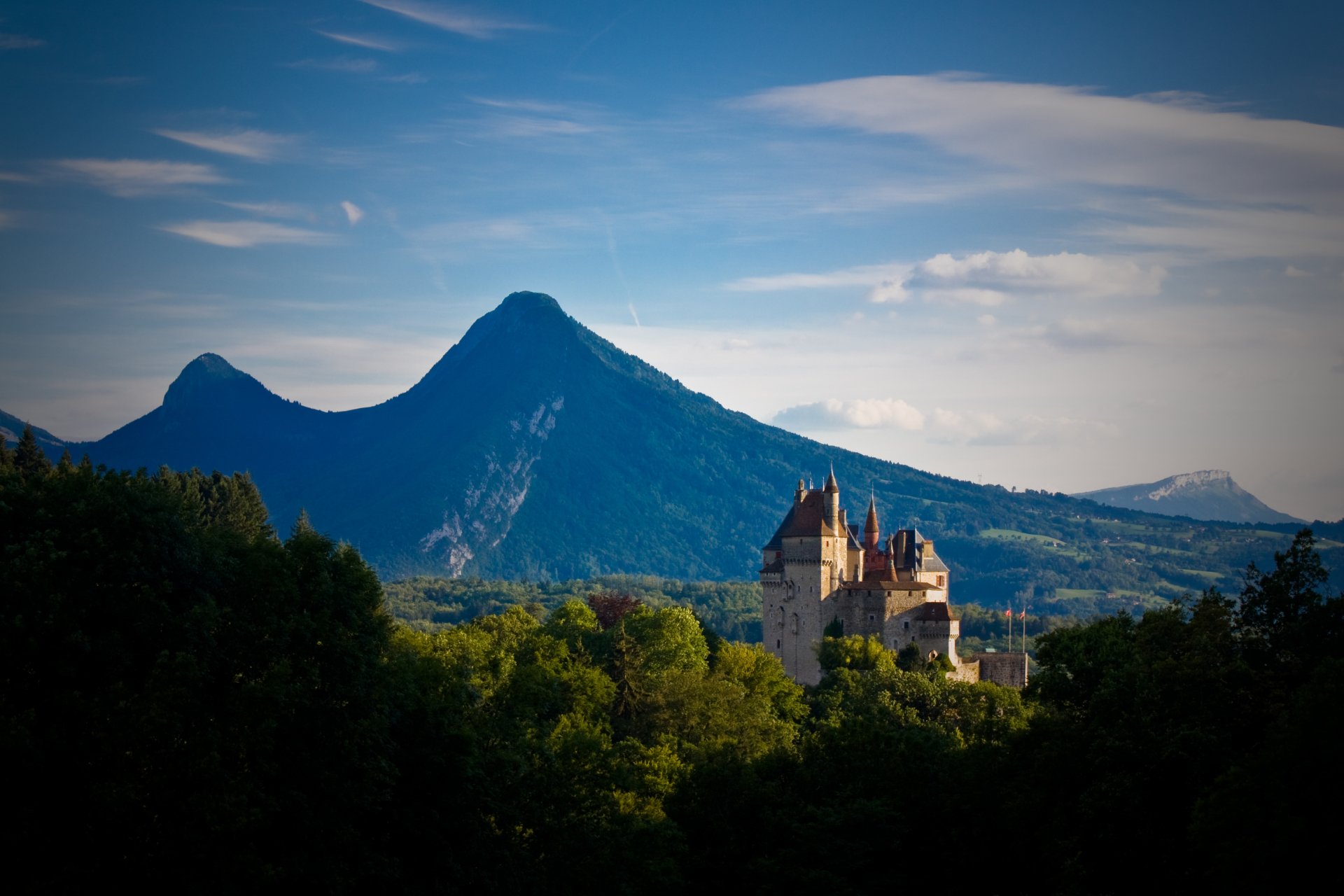 4K Ultra HD PC desktop wallpaper of Chateau de Menthon St. Bernard — a man-made castle perched above forested hills with mountain peaks and a wide blue sky.