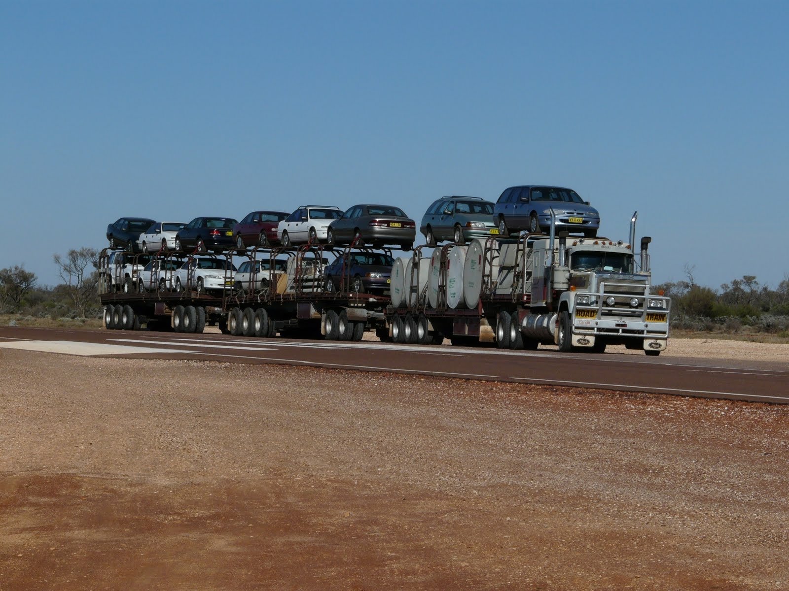 HD PC desktop wallpaper: Mack Trucks car carrier vehicle hauling two trailers loaded with cars across a remote highway under clear blue sky.