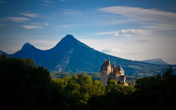 4K Ultra HD PC desktop wallpaper of Chateau de Menthon St. Bernard — a man-made castle perched above forested hills with mountain peaks and a wide blue sky.