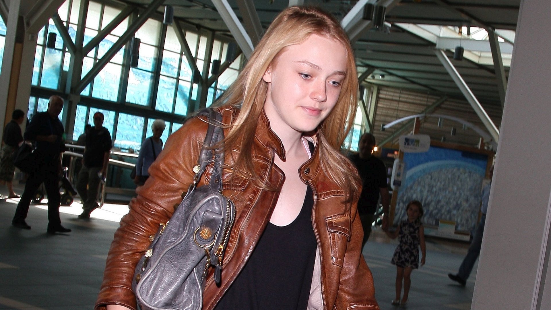 HD PC desktop wallpaper: young blonde woman in a brown leather jacket carrying a gray bag, walking through a sunlit train station or transit terminal.