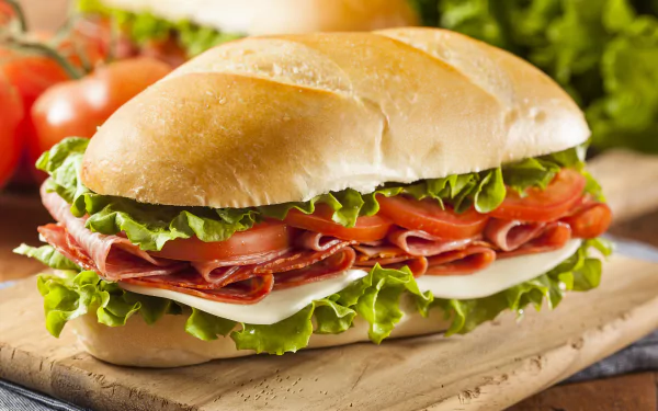 HD desktop wallpaper featuring a fresh lunch sandwich with salami, lettuce, tomato, and cheese on a bread roll, accompanied by salad ingredients in the background.