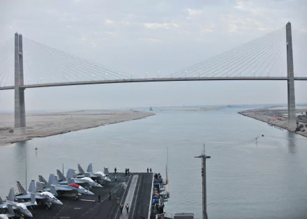 HD desktop wallpaper showing the USS Enterprise (CVN-65) aircraft carrier with jet fighters on deck, sailing under a large cable-stayed bridge in a calm naval setting.