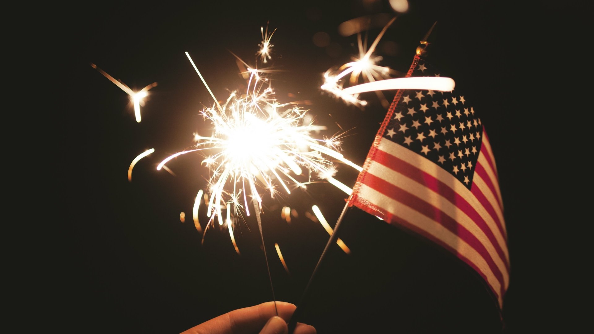 A vibrant 4K Ultra HD desktop wallpaper showing a hand holding a sparkler and an American flag, celebrating the 4th of July Independence Day holiday with fireworks.