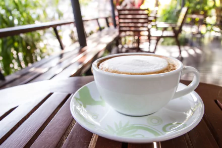 A white cup of frothy coffee sits on a saucer atop a wooden table in an outdoor café setting, captured in 4K Ultra HD quality for a PC desktop wallpaper.