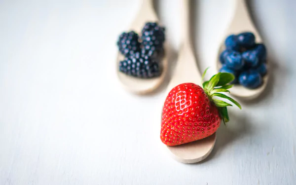 8K Ultra HD PC desktop wallpaper: fresh strawberry on a wooden spoon in foreground, blackberries and blueberries on spoons blurred behind — vibrant berry food close-up.