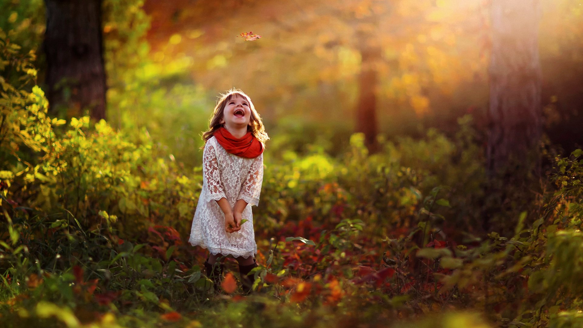 Bright 2K Quad HD PC desktop wallpaper and background photograph of a laughing child in a sunlit forest clearing, leaves drifting in warm autumnal light.
