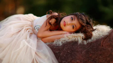A high-definition wallpaper of a child lying on her side, donning a soft, flowing dress, with an expression of serene contemplation against a blurred natural background.