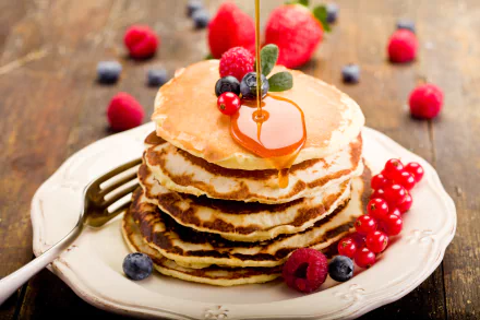 Stacked pancakes drizzled with honey, topped with fresh raspberries, blueberries, and currants. A vibrant and delicious breakfast scene on a rustic wooden table.