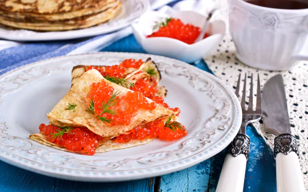 Close-up of delicate crêpes topped with vibrant red caviar and fresh dill on a white plate, set against a rustic blue wooden table, captured in 4K Ultra HD resolution.