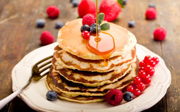 Stacked pancakes drizzled with honey, topped with fresh raspberries, blueberries, and currants. A vibrant and delicious breakfast scene on a rustic wooden table.