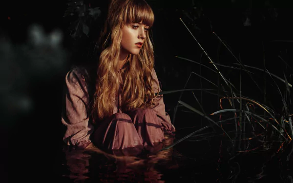 Blonde woman seated in dark water among reeds, moody lighting and reflective surface — 5K Ultra HD PC desktop wallpaper and background.