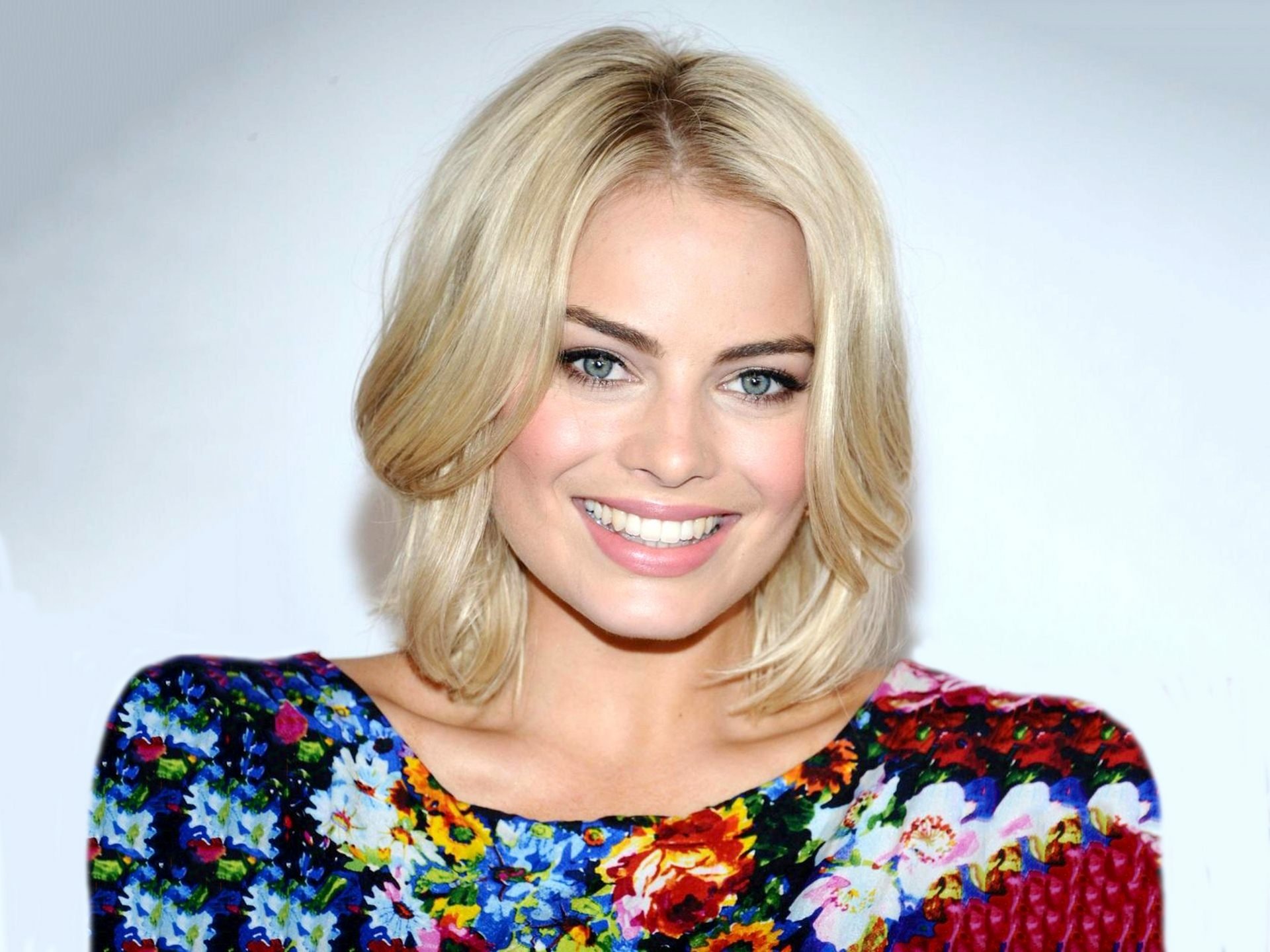HD desktop wallpaper featuring Margot Robbie smiling confidently, dressed in a vibrant floral top, showcasing her stylish presence and captivating blue eyes.