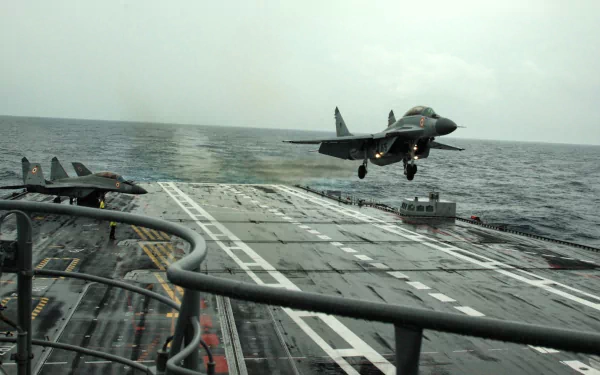 HD desktop wallpaper showing the INS Vikramaditya (R33) aircraft carrier with a military jet landing on its deck amid a calm sea and cloudy sky.