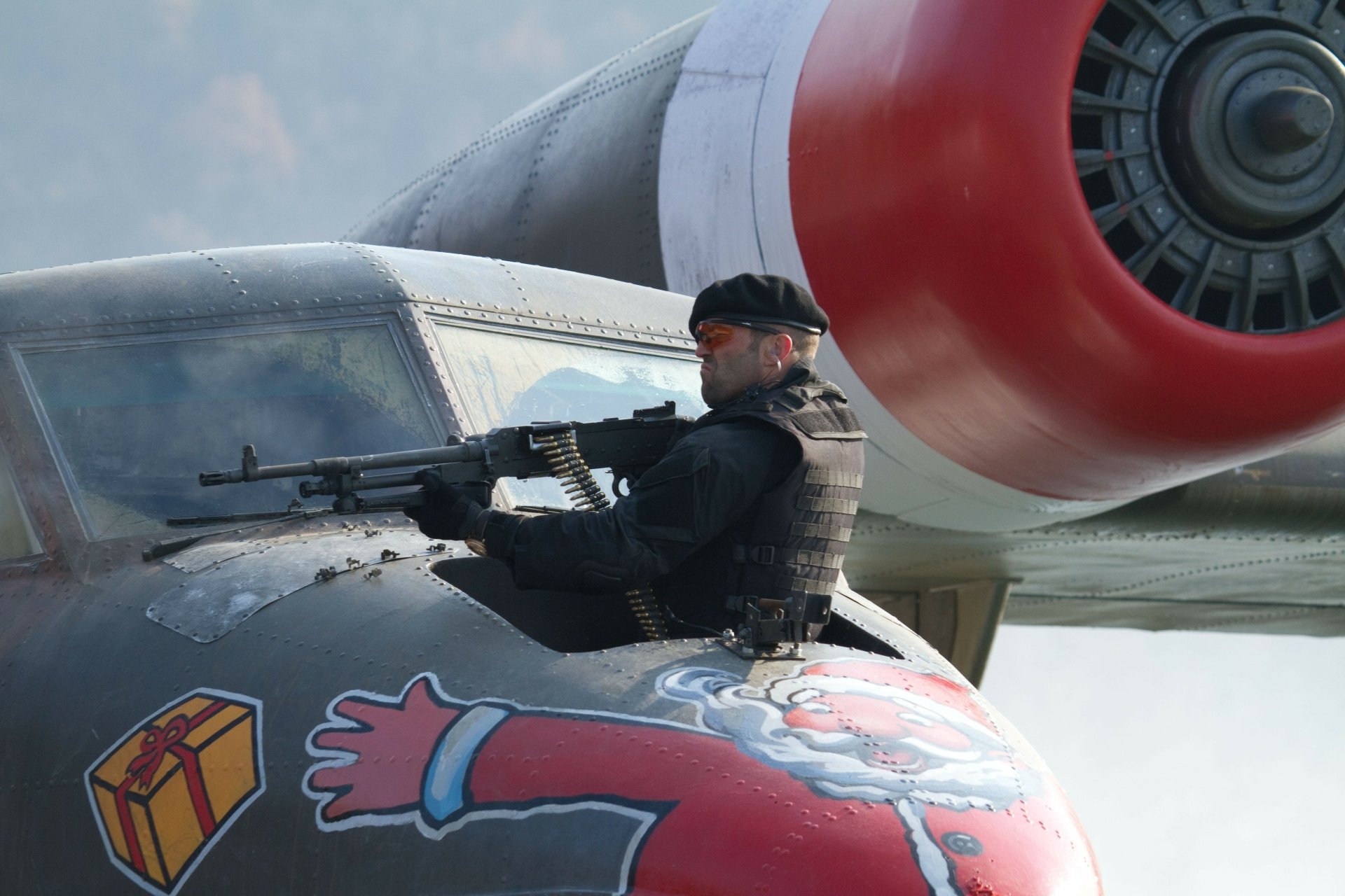 5K Ultra HD PC desktop wallpaper of an action-movie mercenary on a painted bomber nose, firing a mounted machine gun — evokes The Expendables 2.