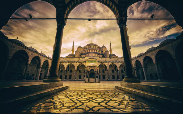 Religious view of the Sultan Ahmed Mosque courtyard framed by arches, ornate patterned floor and dramatic golden sky — HD PC desktop wallpaper and background.