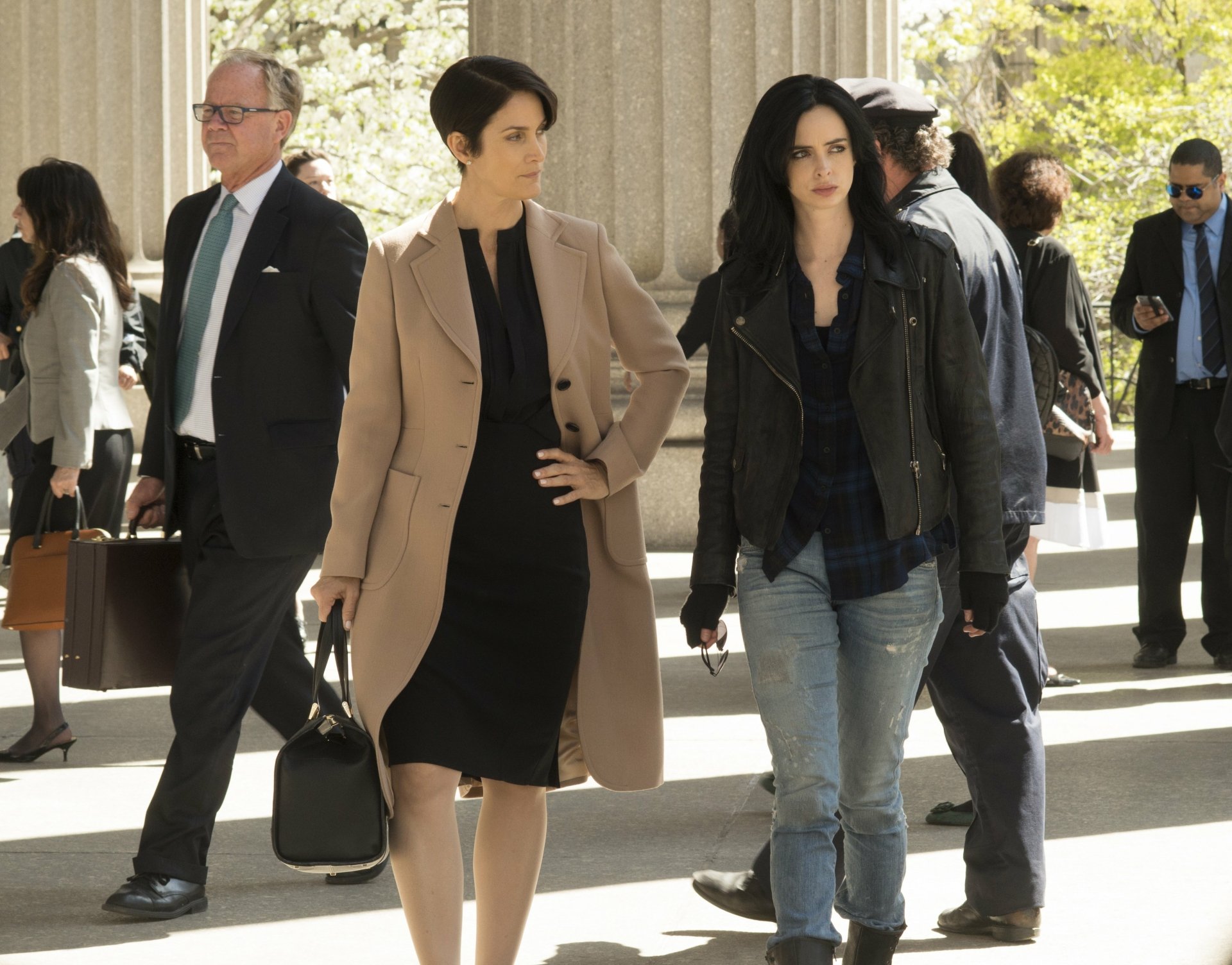 2K Quad HD PC wallpaper: Jessica Jones TV scene of two women walking through a courthouse plaza — one in a beige trench and dress with handbag, the other in a black leather jacket and jeans.