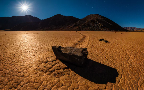 HD PC desktop wallpaper: Death Valley nature landscape — cracked desert floor with a single stone casting a long shadow under a bright sun and dark silhouetted mountains.
