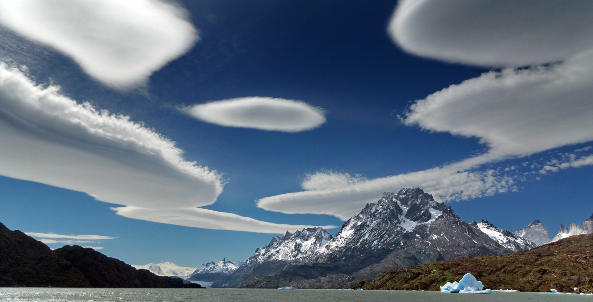 A stunning Patagonian landscape featuring a tranquil lake, majestic mountains, and floating icebergs, under a vibrant blue sky with unique cloud formations.