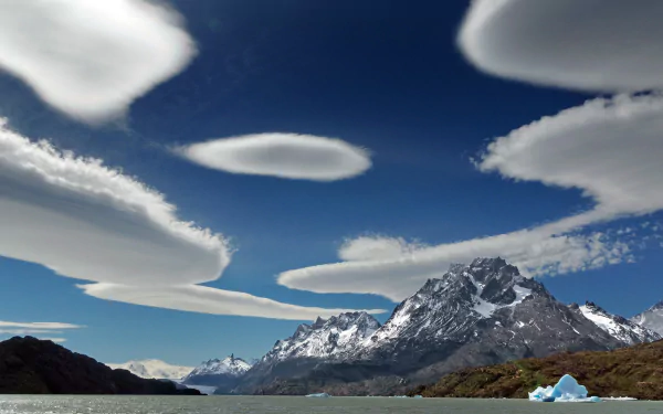 A stunning Patagonian landscape featuring a tranquil lake, majestic mountains, and floating icebergs, under a vibrant blue sky with unique cloud formations.