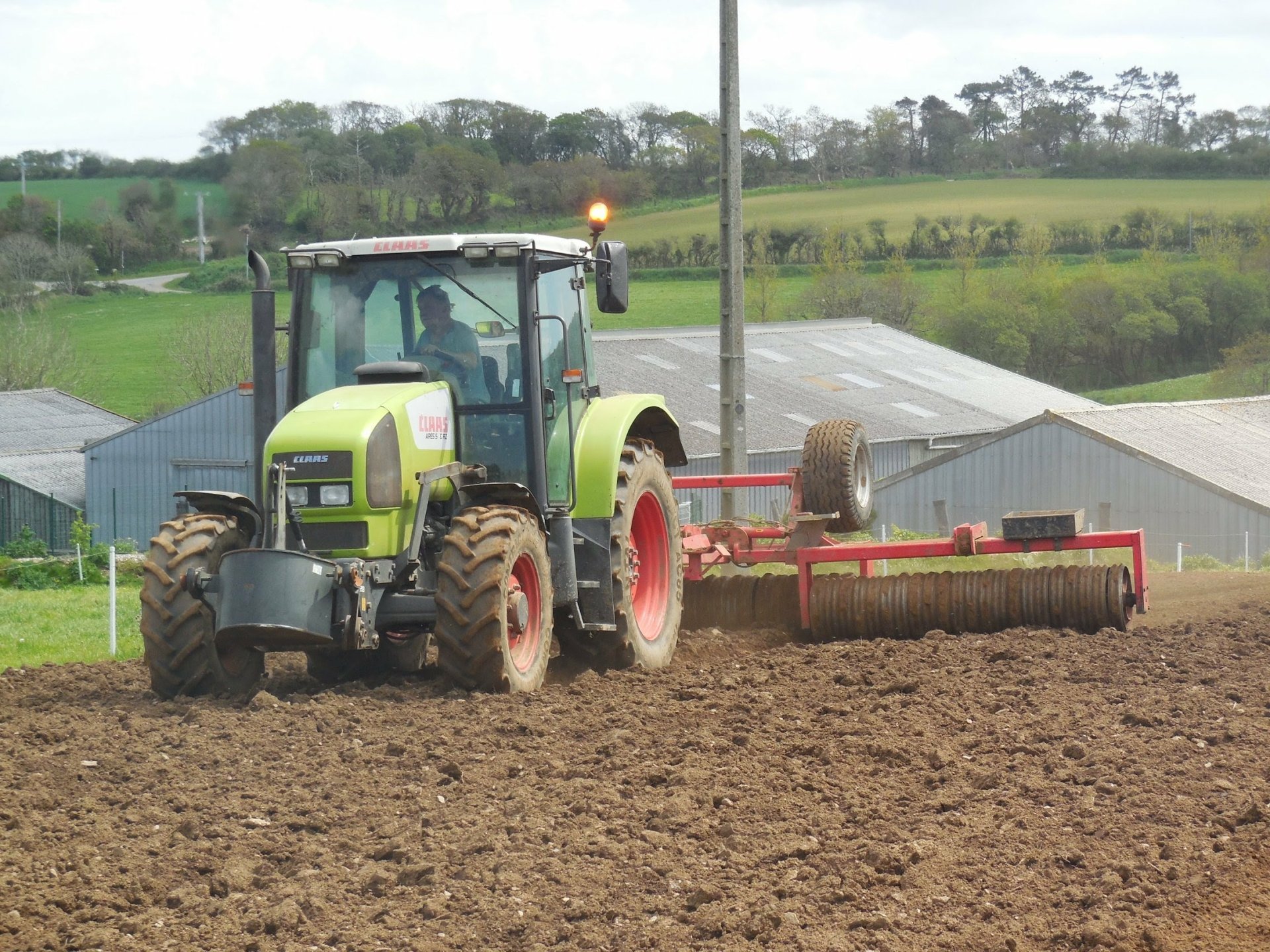 HD PC desktop wallpaper featuring a Claas tractor working on a plowed field in a rural setting with farm buildings and greenery in the background.