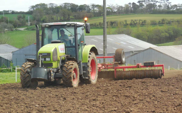 HD PC desktop wallpaper featuring a Claas tractor working on a plowed field in a rural setting with farm buildings and greenery in the background.