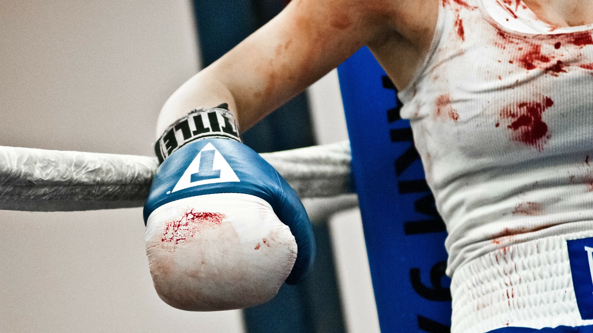 HD PC desktop wallpaper showing a close-up of a bloodied boxing glove and arm resting on the ring ropes during a sports match.
