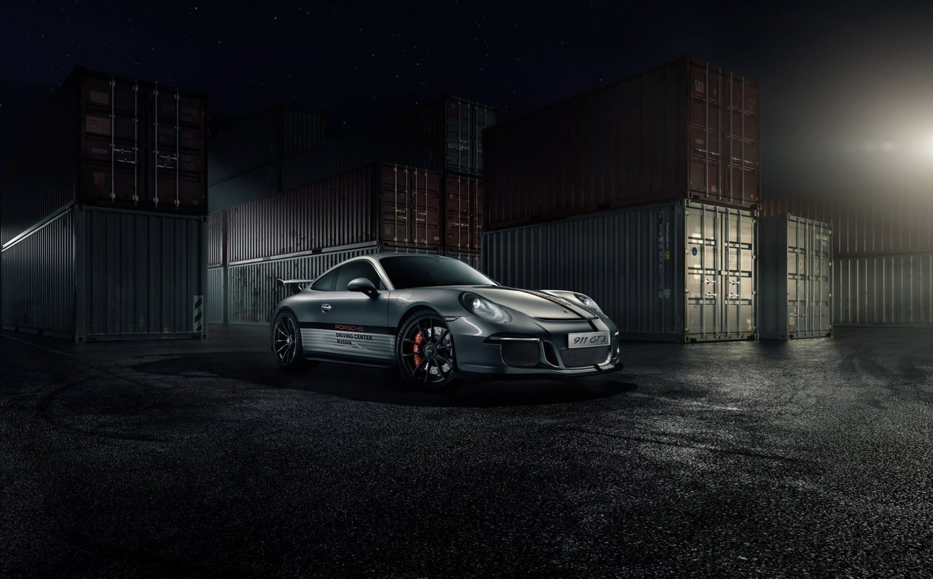 Silver Porsche 911 GT3 supercar parked on wet ground with stacked shipping containers in the background, captured in a high-definition desktop wallpaper style.