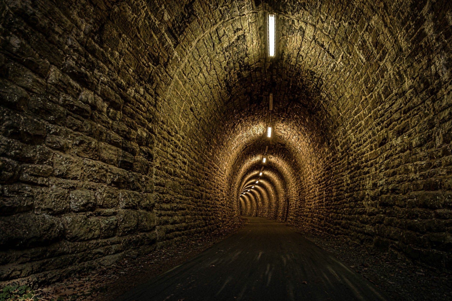 4K Ultra HD wallpaper of a man-made stone tunnel illuminated by evenly spaced overhead lights, showcasing textured brick walls and a vanishing point in the distance.