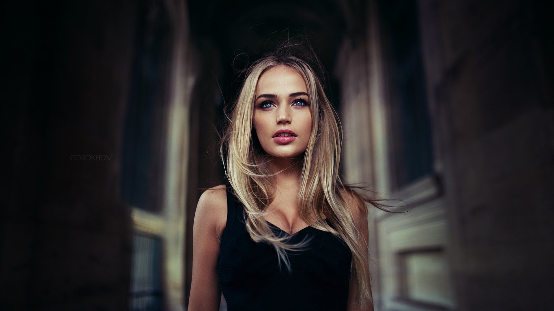 A high-definition desktop wallpaper of a woman model with long blonde hair and a black top, standing in a dimly lit, elegant corridor.