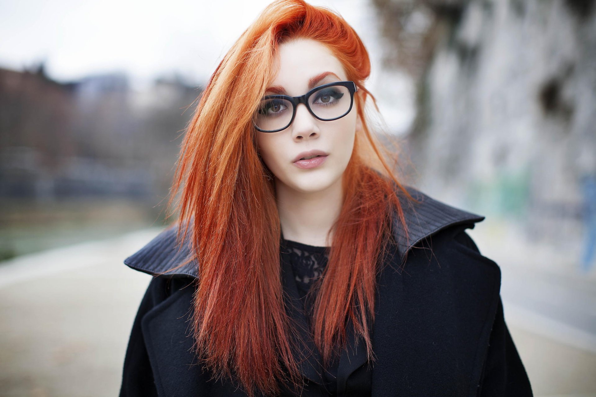 A woman with long red hair and brown eyes wears glasses outdoors, surrounded by a soft bokeh background, making for an engaging HD desktop wallpaper.