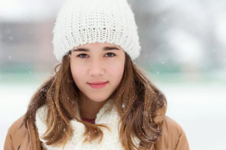 Smiling brunette woman with brown eyes wearing a white knit hat and scarf stands outdoors during a gentle winter snowfall, captured in HD for a desktop wallpaper.