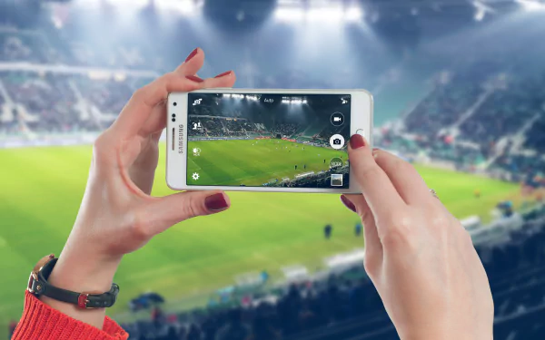 Hands holding a Samsung phone capture a soccer match in a vibrant 4K Ultra HD stadium, showcasing a man-made digital moment in sports photography.