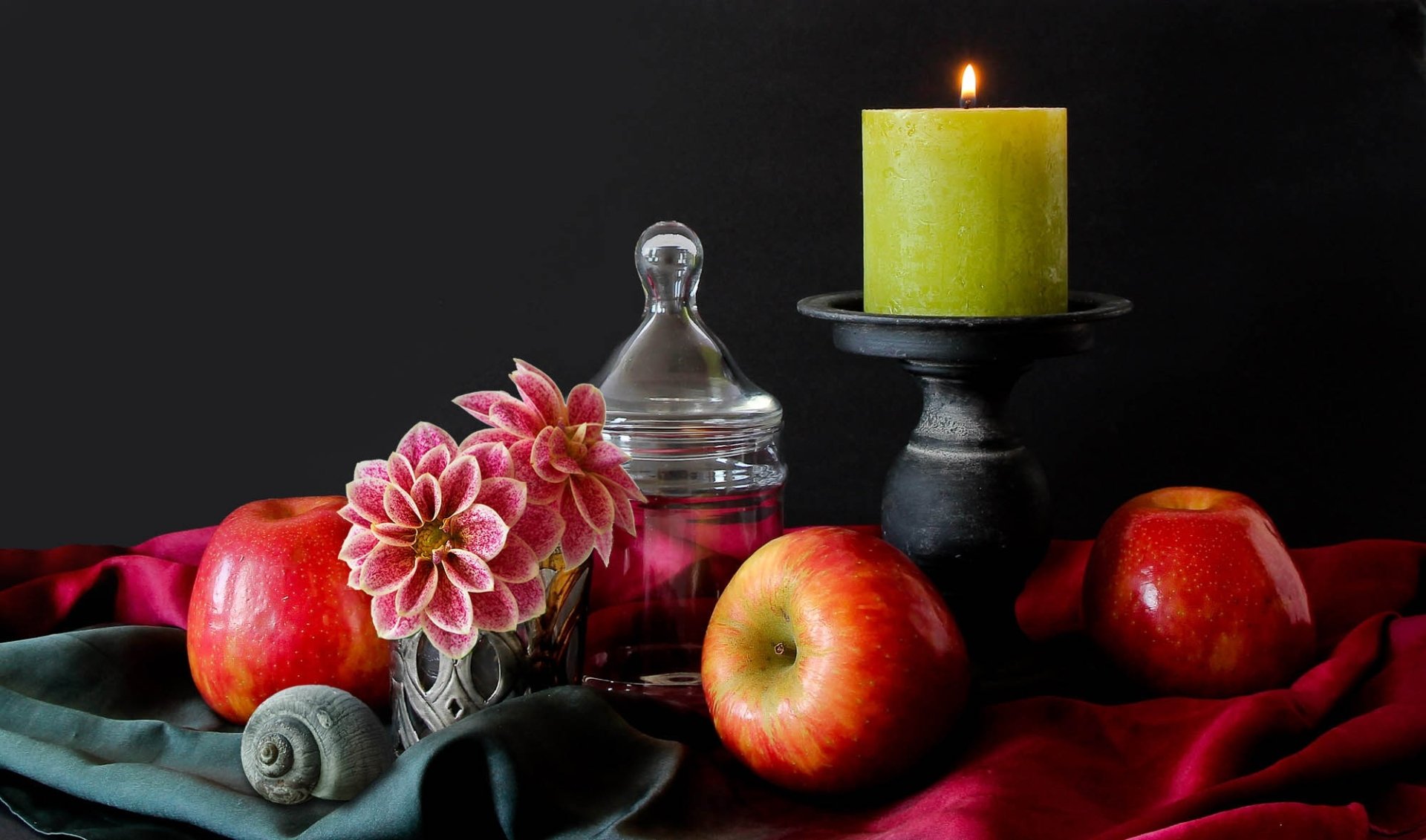 HD still-life photography for PC desktop wallpaper: red apples, pink flowers, a lit green candle on a black stand, glass jar and draped fabric against a dark background.