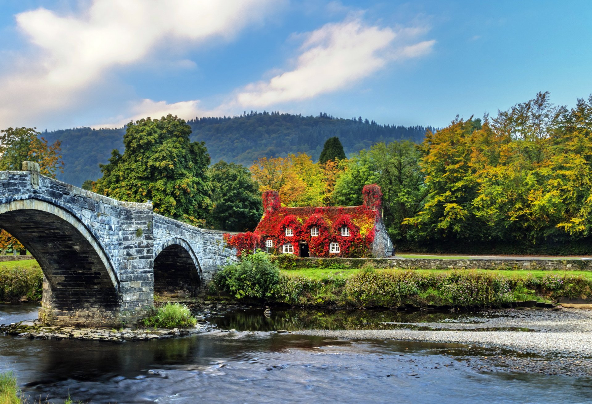 A picturesque fall scene in Wales featuring a historic stone bridge over a river and a charming red ivy-covered house surrounded by autumn foliage.