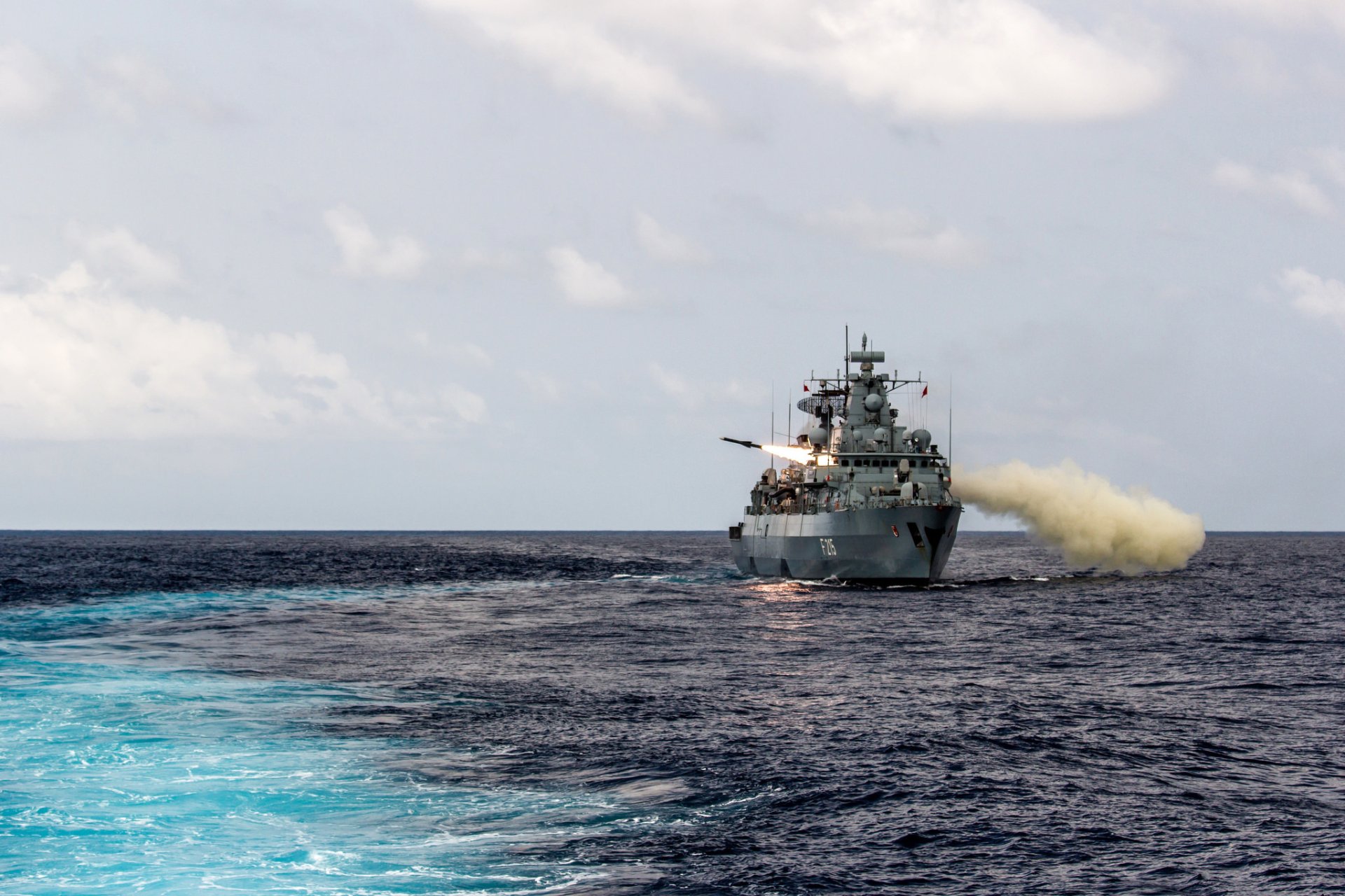 HD PC desktop wallpaper: German Navy frigate Brandenburg (F 215) warship firing at sea, military vessel against an open ocean background.