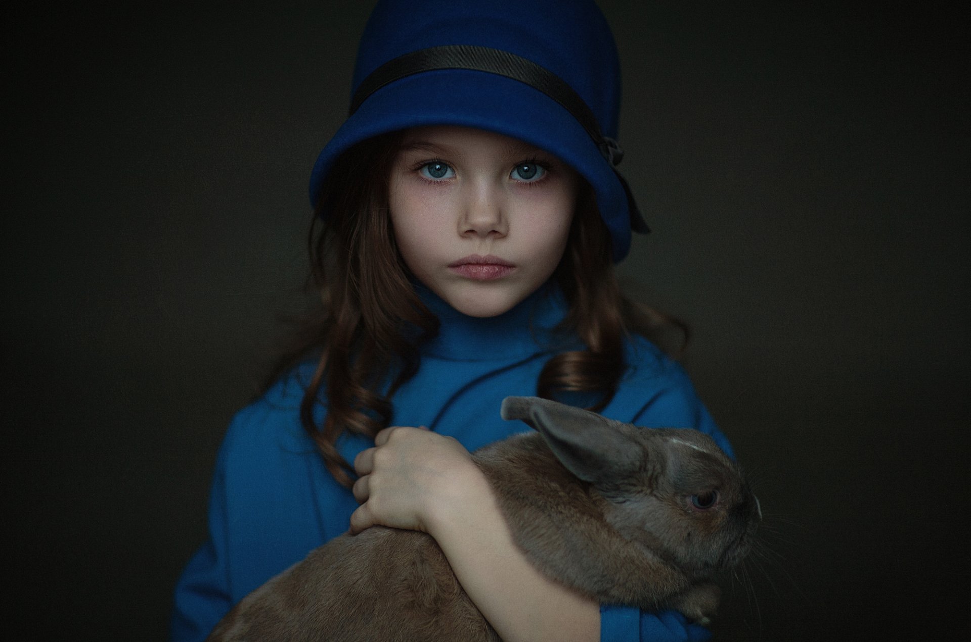 A portrait of a child with blue eyes and brunette hair, wearing a blue hat and coat, holding a rabbit. The dark background enhances the focus on the child and rabbit. HD desktop wallpaper and background.