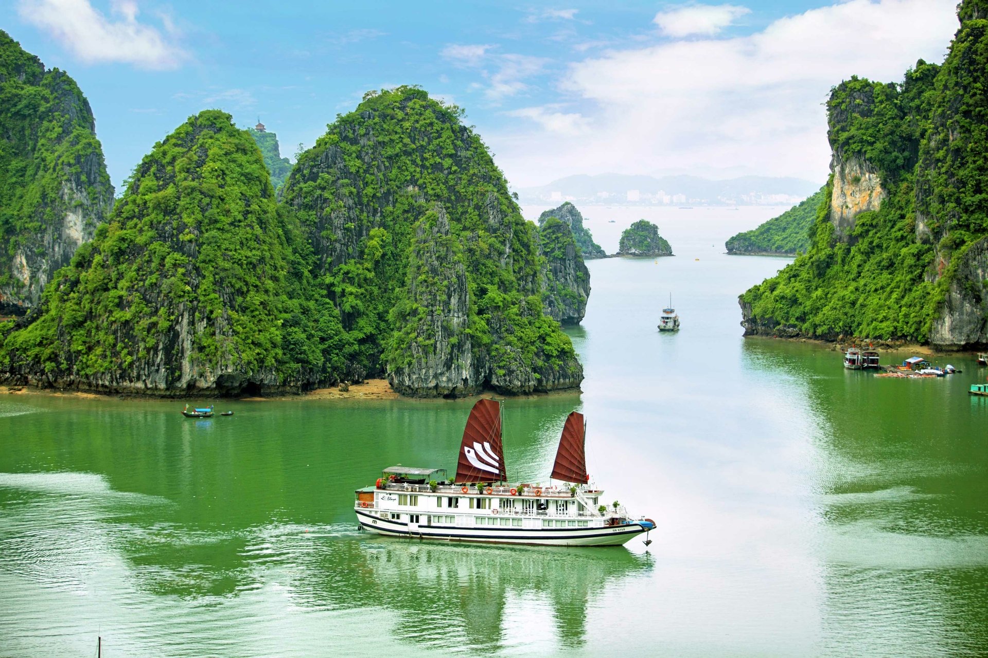 HD desktop wallpaper of a scenic Vietnam bay featuring lush green limestone islands and a traditional boat cruising the calm, emerald waters.
