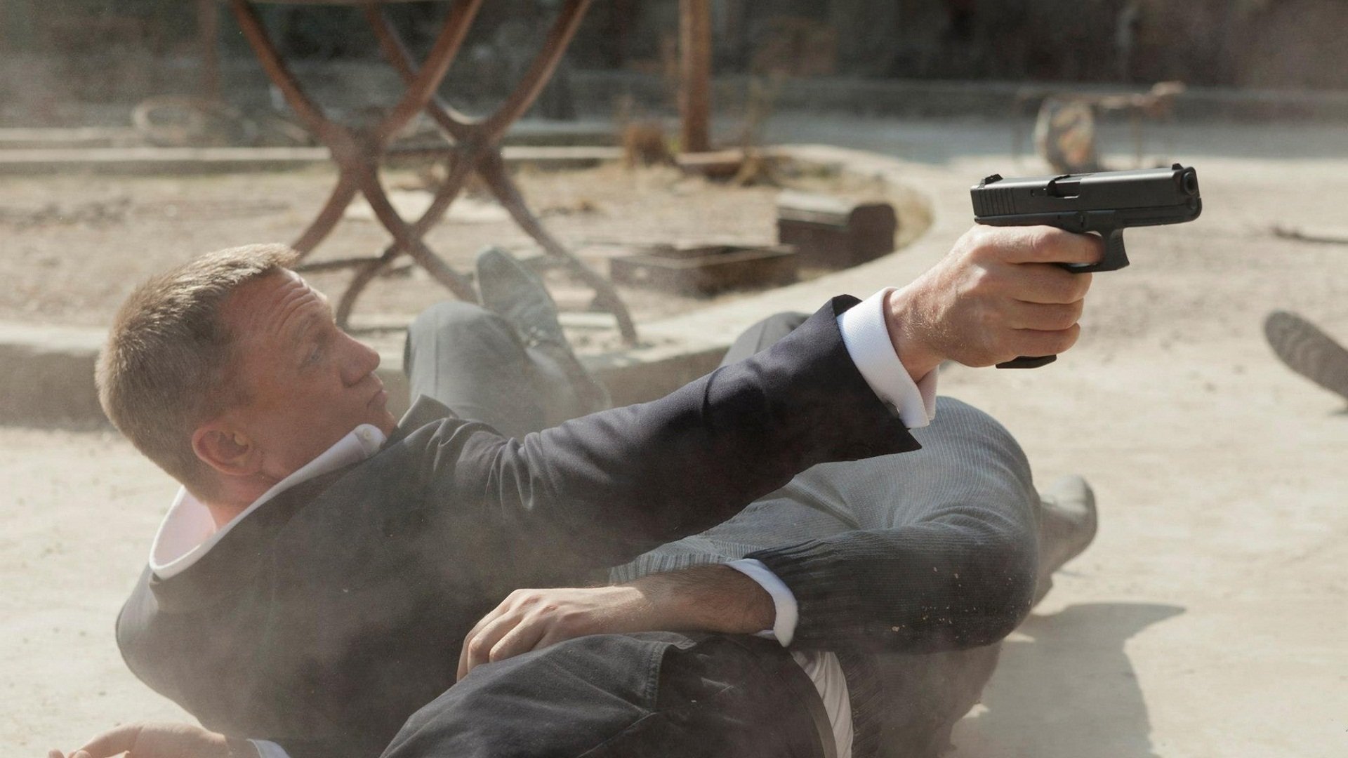 HD PC desktop wallpaper: cinematic Skyfall movie scene — suited spy sprawled on the ground firing a handgun in a sunlit outdoor café.