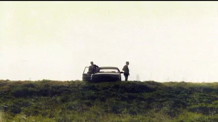 HD PC desktop wallpaper featuring a scene from the TV show True Detective, with two figures standing near a car on a grassy hill under a pale sky.