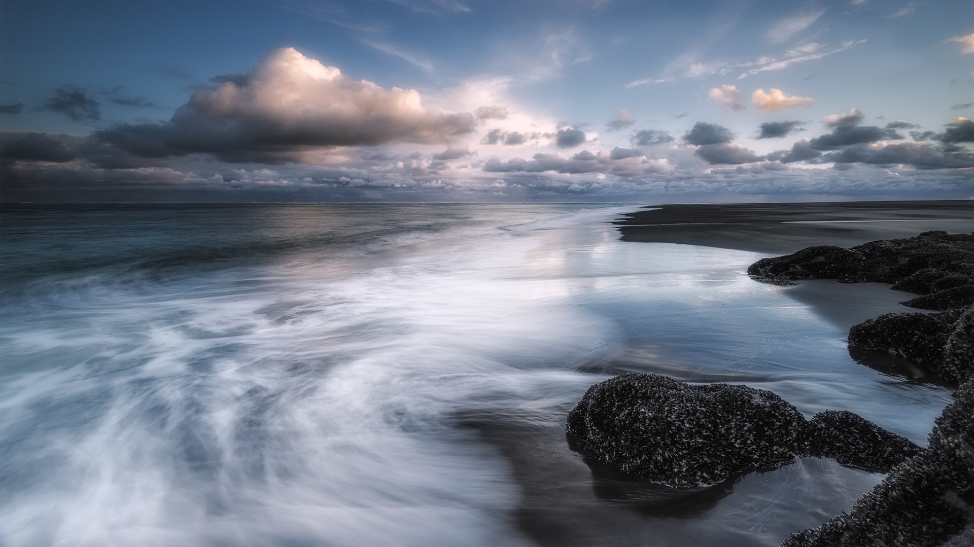 HD PC desktop wallpaper/background: tranquil ocean beach at low tide with clouds on the horizon, smooth long-exposure waves washing around dark rocks — serene coastal nature scene.