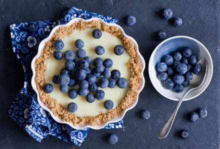 A freshly baked tart topped with blueberries, accompanied by a bowl of blueberries and a spoon, set on a dark surface with a blue patterned cloth.