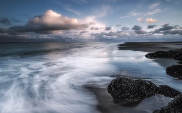 HD PC desktop wallpaper/background: tranquil ocean beach at low tide with clouds on the horizon, smooth long-exposure waves washing around dark rocks — serene coastal nature scene.