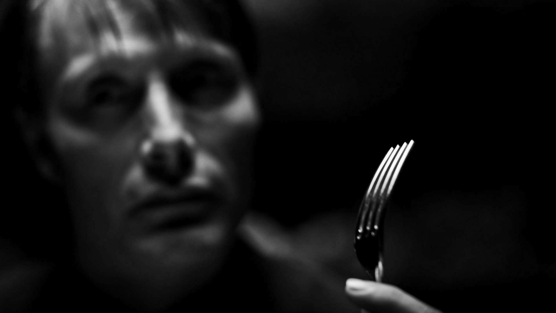 HD desktop wallpaper featuring a blurred close-up of Hannibal from the TV show, holding a fork against a dark background.