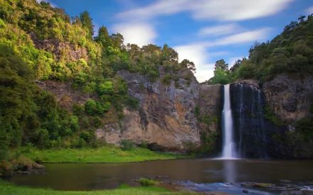 A beautiful HD wallpaper showcasing Hunua Falls in New Zealand. The serene scene includes a waterfall cascading down a cliff into a river, surrounded by lush greenery and under a vibrant blue sky.