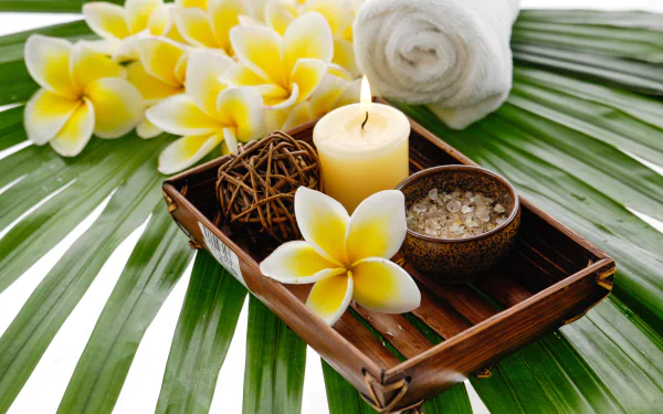 A 4K Ultra HD still life spa scene featuring frangipani flowers, a lit candle, bath salts, and a rolled white towel arranged on a wooden tray atop green palm leaves.